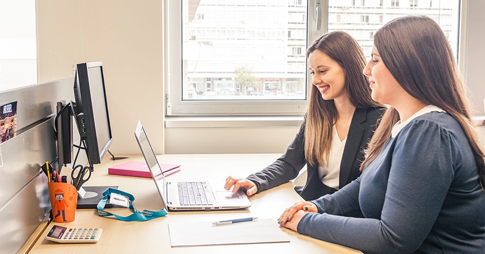 Two smiling women at an office desk looking at a laptop screen (photo)