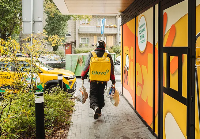 A delivery driver with a Glovo backpack carrying several bags while walking along a Biek store photographed from behind (photo)