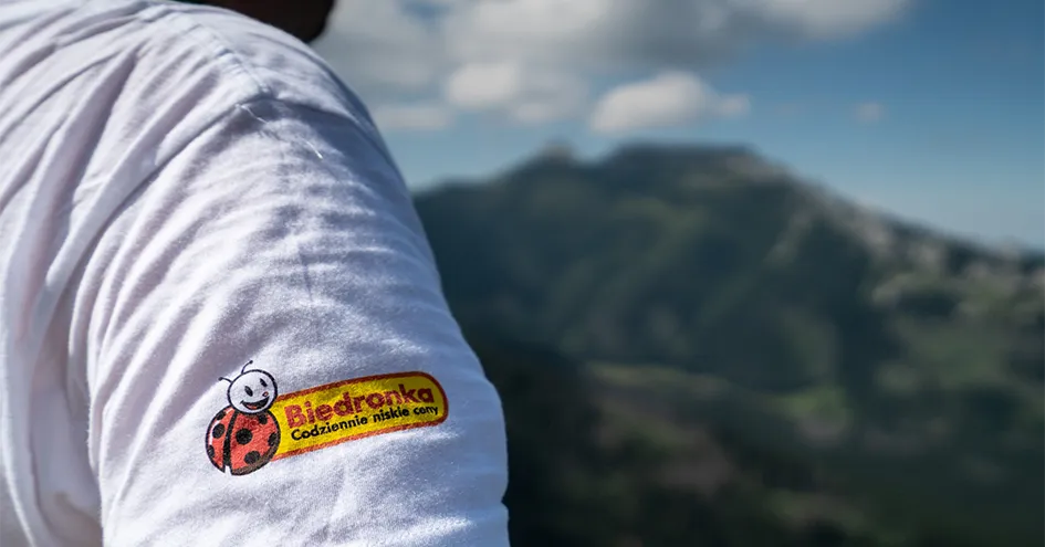 A close-up of a person’s shoulder, with a white T-shirt bearing a Biedronka logo, against a backdrop of a mountain. (photo)
