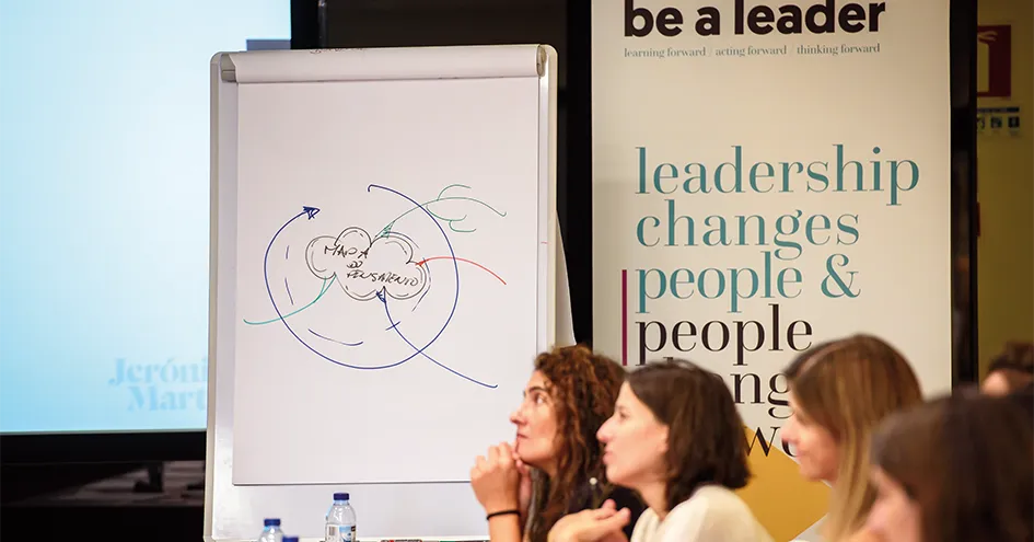 Photo of a lecture. Focus is on the flipchart showing some scribbles and a banner in the background (photo)