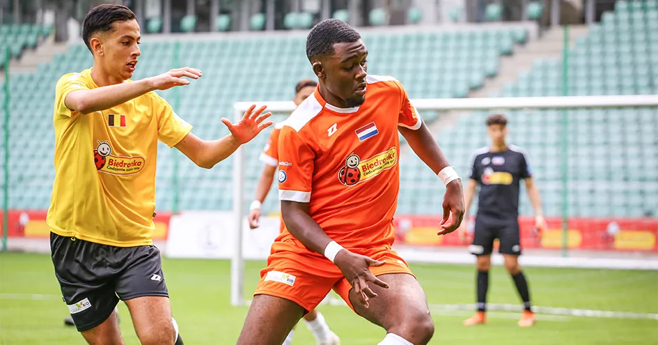 Two opposing players in a football game. Both jersies have the Biedronka logo. The goal and goalkeeper are blurred out in the background.