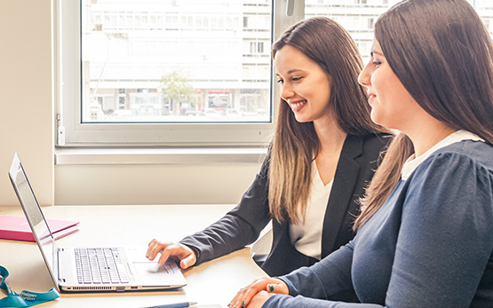 Duas mulheres sorridentes numa secretária de escritório a olhar para o ecrã de um computador portátil (foto)