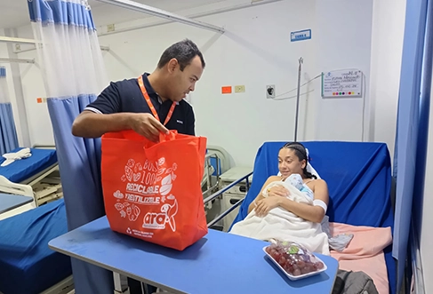 Uma mulher com um recém-nascido numa cama de hospital recebe um saco vermelho cheio de compras entregue por um homem. (foto)