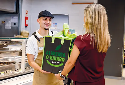 Um colaborador do Pingo Doce a entregar um saco reutilizável grande onde está escrito "Alimenta o Bairro" a uma cliente em frente a um balcão (foto)
