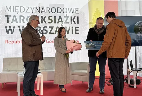 Uma mulher e dois homens a entregarem prémios (um cheque e um ramo de flores) ao vencedor numa cerimónia de entrega de prémios (foto)