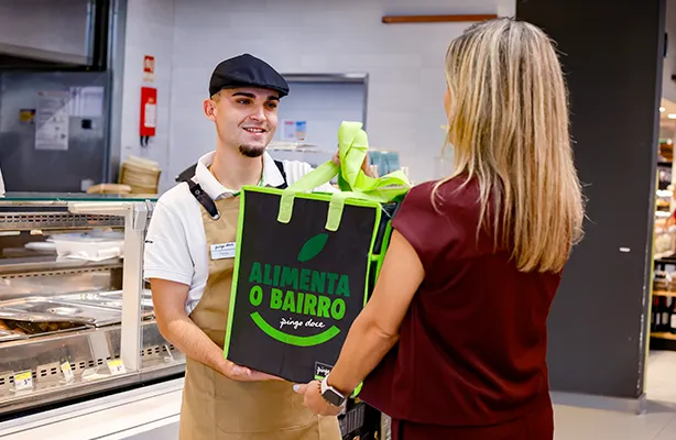 Um colaborador do Pingo Doce a entregar um saco reutilizável grande onde está escrito "Alimenta o Bairro" a uma cliente em frente a um balcão (foto)
