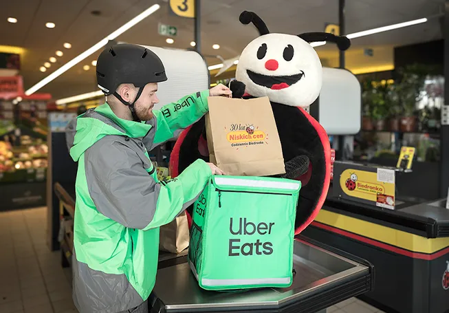 Um motorista de entregas da Uber Eats a colocar um saco de papel na sua mochila na caixa de pagamento, com um grande peluche da Biedronka atrás dele (foto)