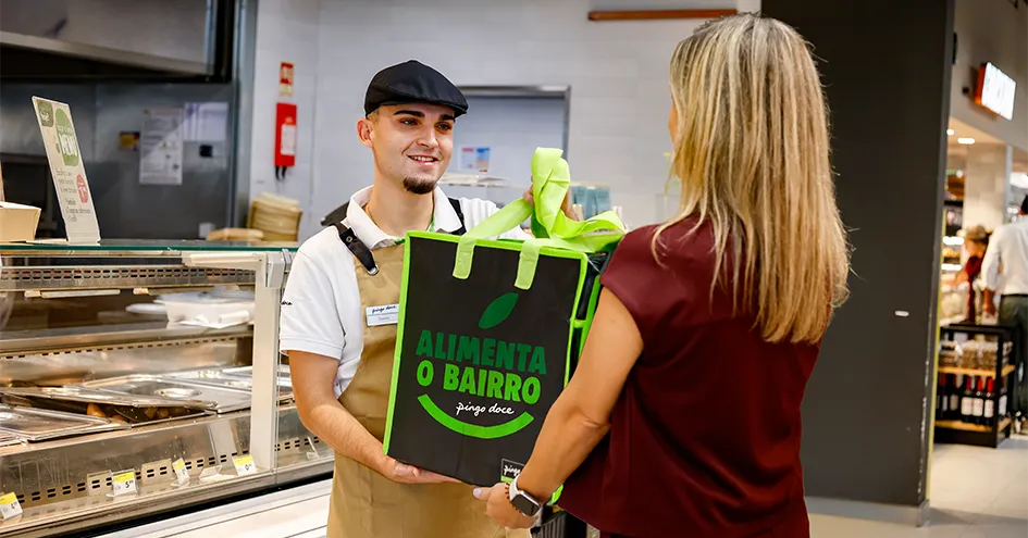 Um colaborador do Pingo Doce a entregar um saco reutilizável grande onde está escrito "Alimenta o Bairro" a uma cliente em frente a um balcão (foto)