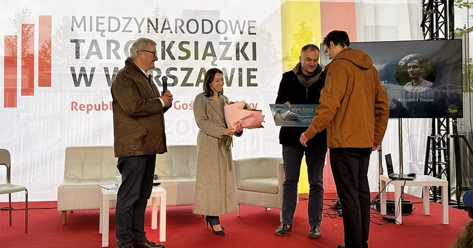 Uma mulher e dois homens a entregarem prémios (um cheque e um ramo de flores) ao vencedor numa cerimónia de entrega de prémios (foto)
